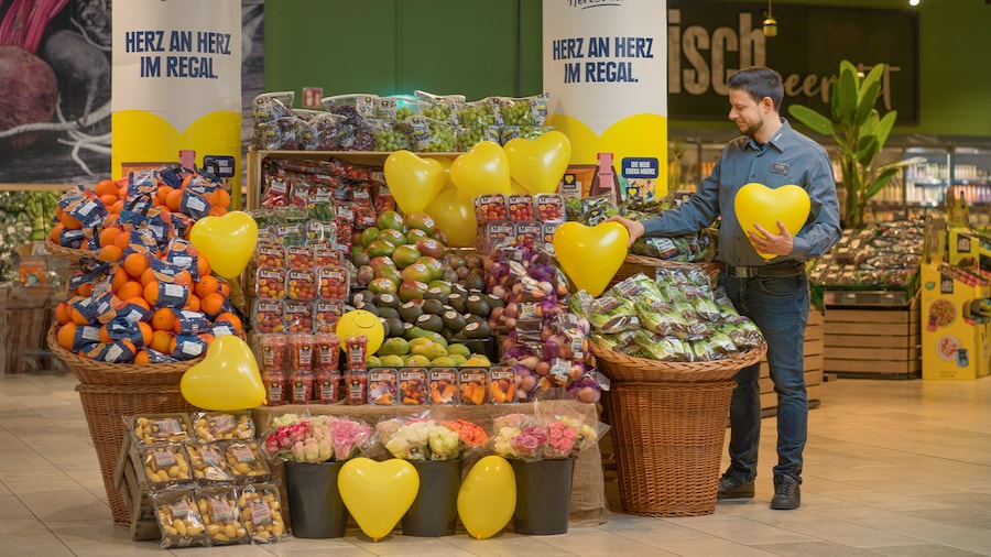 Verkäufer arrangiert Obst und Gemüse mit gelben Herz-Luftballons im Supermarkt.