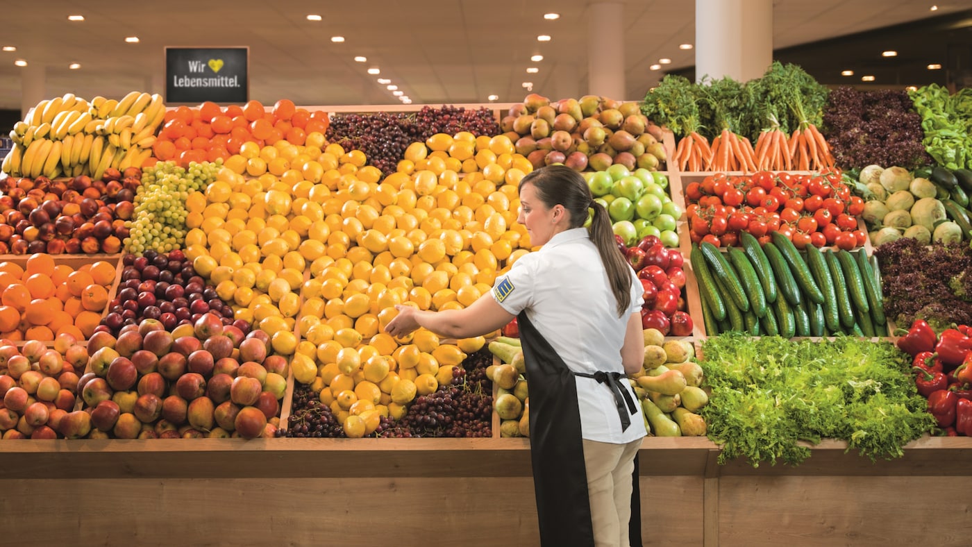 Obstabteilung im Markt: Im schrägen Regal liegen viele gelbe Zitronen zu einem großen Herz, daneben Obst, davor eine Frau.
