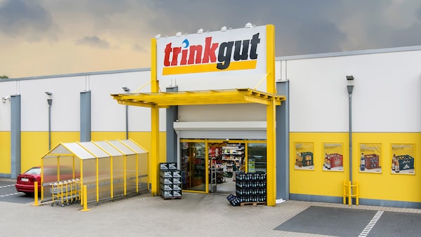 Außenansicht: Markteingang Trinkgut-Getränkemarkt, Getränkekisten daneben und großes Logo über dem überdachten Eingang.